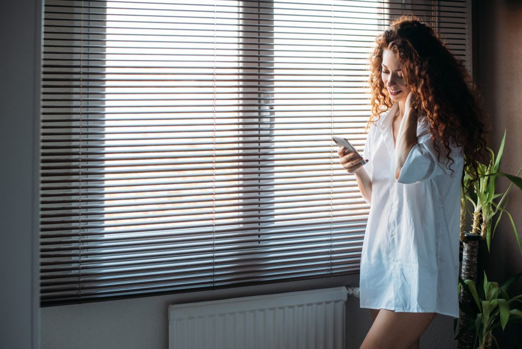 Beautiful woman standing by a roller blind with mobile phone