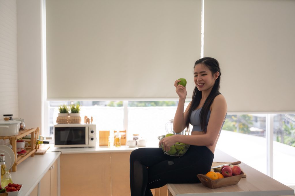 young Asian woman cooking vegetable healthy food and eating or drinking in home kitchen front of moisture resistant roller blinds